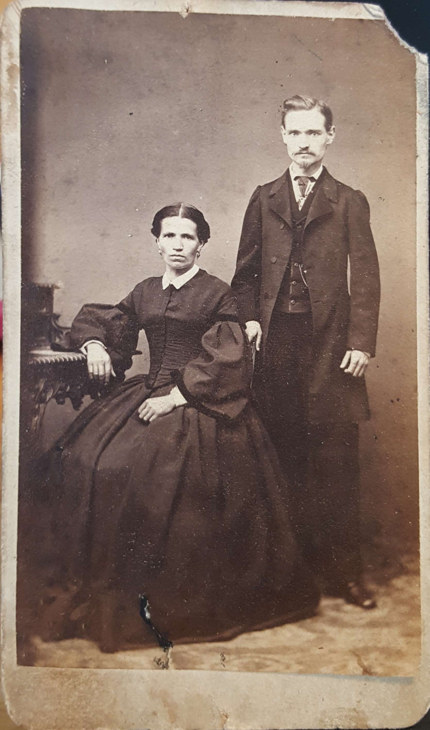 A carte de visite of a seated woman and a standing man, both in dark clothing, photographed in a studio against a plain backdrop. She rests one arm on a carved table; he stands beside her with a hand at her shoulder. The card's mount is worn at the corners and stained along the lower edge.