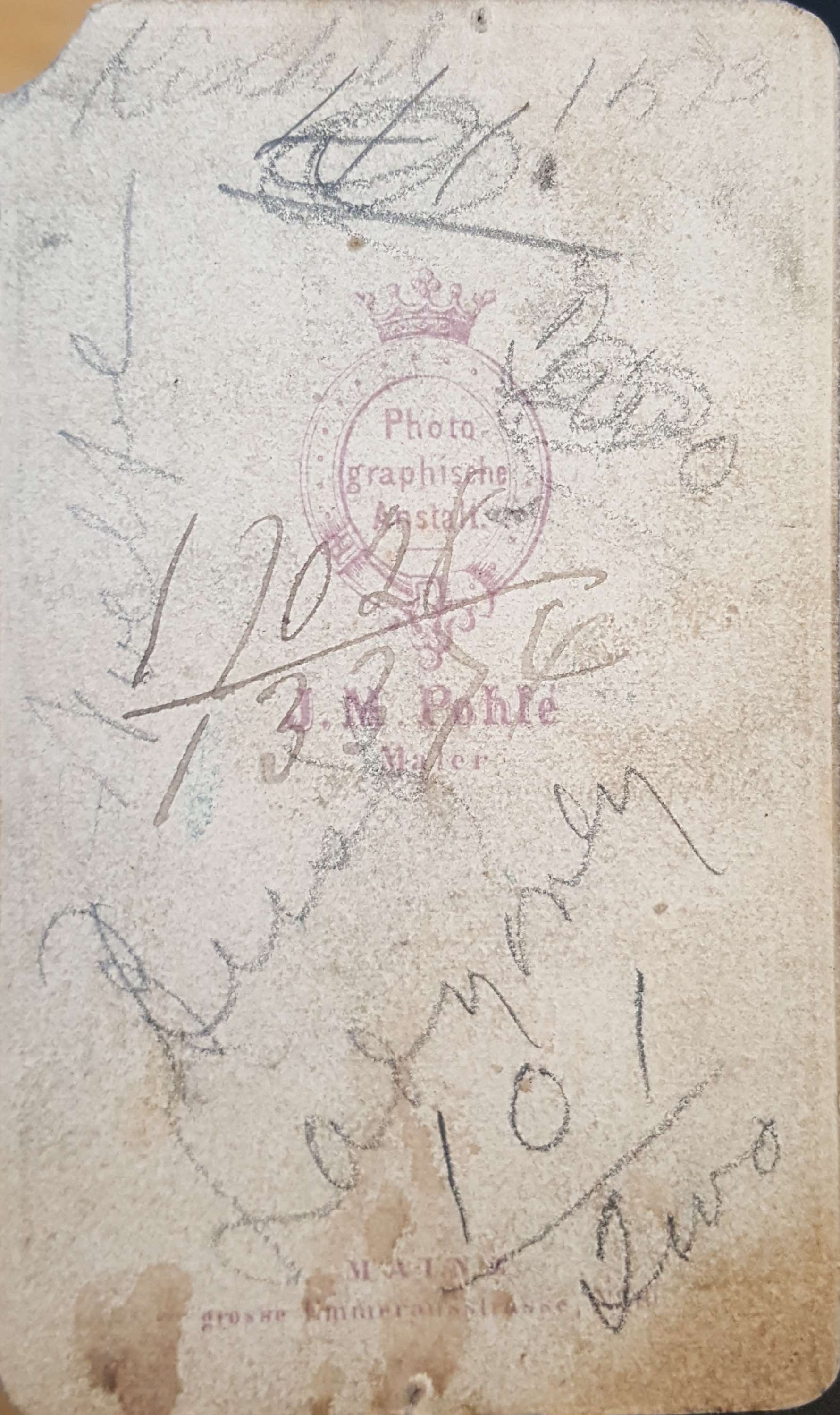 The back of the same card, worn and foxed. A photographer's printed emblem sits in the middle — a crown above an oval reading Photographische Anstalt, and beneath it the name J. B. Pohle, Mainz. Across and around it, several different hands have written in pencil: names, numbers and what may be dates, overlapping each other and mostly illegible.