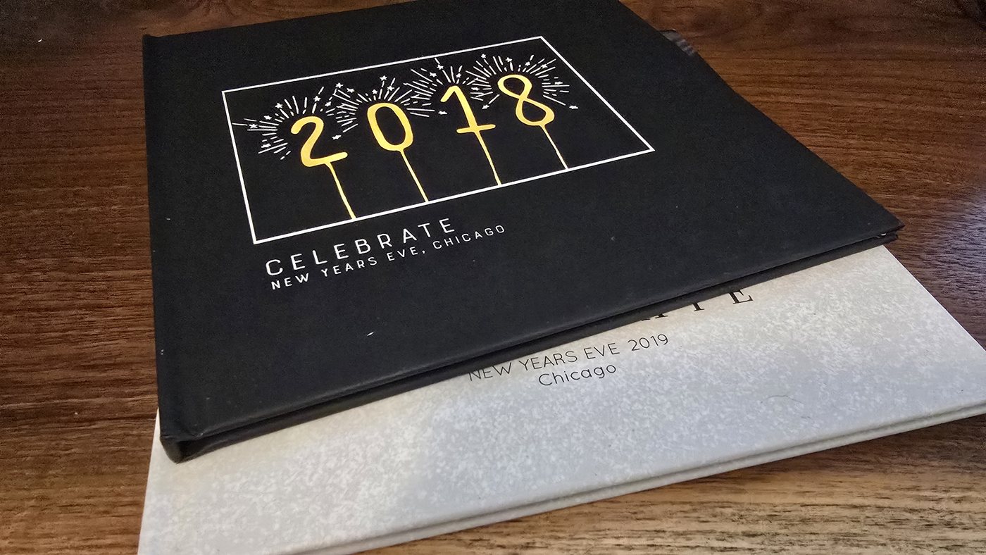 Two Milestone books stacked on a wood table. The top volume is black cloth, blocked in white and gold with sparkler numerals reading 2018 and the words Celebrate, New Years Eve, Chicago. Beneath it a pale grey volume is blind-blocked New Years Eve 2019, Chicago.